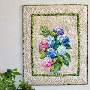May include: A rectangular fabric wall hanging featuring a central applique of blue, pink, and white hydrangea flowers in a vase. The quilt has a beige background with a stippled texture, framed by a green patterned border and a beige outer border.