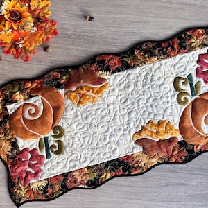 May include: A quilted table runner with a white center and a floral and leaf border. The runner features appliqued pumpkins and leaves in shades of orange, red, and yellow. The runner is approximately 30 inches long.