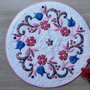 Può includere: Un sottopentola rotondo e trapuntato bianco con un bordo rosa. Il sottopentola presenta un motivo floreale con fiori rosa e blu, accenti paisley e un fiore centrale. Tulipani rossi sono nell'angolo in basso a destra.