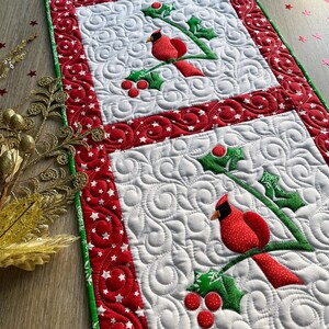 May include: A rectangular quilted table runner featuring three white square panels, each decorated with an appliqued red cardinal bird perched on green holly branches with red berries, surrounded by a red border with white star patterns.