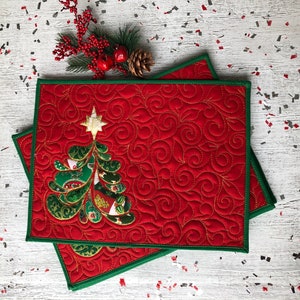 Könnte beinhalten: Zwei rote, gesteppte Tischsets mit grünem Rand. Jedes Tischset hat eine Weihnachtsbaumapplikation mit einem goldenen Stern und dekorativen Nähten. Festliche Weihnachtsdekoration.