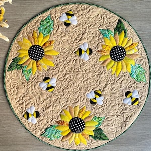 Op de afbeelding: Een ronde, gewatteerde placemat met een beige achtergrond, met gele zonnebloemen met zwarte stippen in het midden en groene bladeren. Verschillende geappliqueerde bijen met zwart-gele strepen zijn verspreid rond de bloemen. De rand heeft een groene rand.