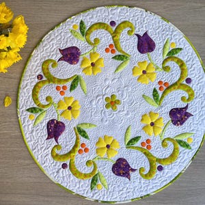 May include: A round, white quilted placemat with a floral design. The design features yellow flowers, purple tulips, green leaves, and orange berries. The placemat has a green border and is placed on a wooden surface.