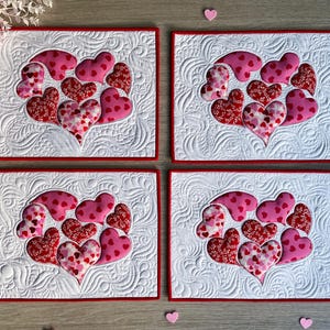 May include: Four Valentine's Day placemats. Each features a white quilted background with a red border. A cluster of pink and red hearts is appliqued on each placemat. The hearts have various patterns, including polka dots and floral designs.