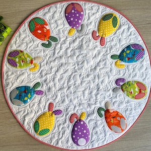 May include: A circular, white quilted Easter placemat with a red trim. It features appliqued Easter egg designs in various colours and patterns, each with bunny ears and feet. A small bouquet of purple flowers is in the corner.