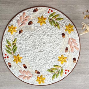 May include: A circular quilted placemat featuring a white background with intricate swirling stitch patterns. A wreath design of yellow flowers, green leaves, pink foliage, small brown acorns, and clusters of red berries borders the center.