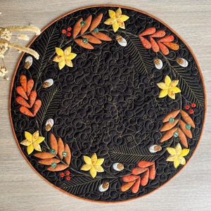 Puede incluir: Un mantel individual redondo y negro acolchado con un diseño decorativo de hojas y flores otoñales. El mantel individual presenta hojas, flores y bayas aplicadas en naranja, amarillo y blanco, con costuras doradas y un borde marrón. El fondo tiene un patrón arremolinado.
