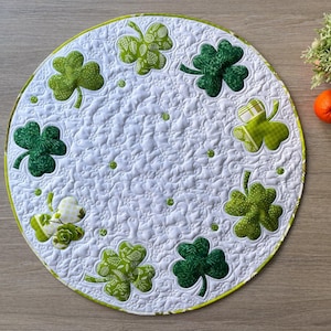 May include: A round, white quilted placemat with a green border. The placemat features eight appliqued shamrocks in various shades of green and patterns. Small green dots are scattered across the white background. A small plant and orange are in the background.