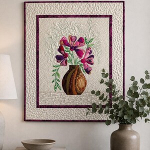 Peut inclure: Une tenture murale rectangulaire en tissu avec un fond crème matelassé et une bordure violet foncé. Le centre présente un vase marron appliqué rempli de fleurs stylisées roses et violettes, agrémenté de feuilles vertes et de coutures.