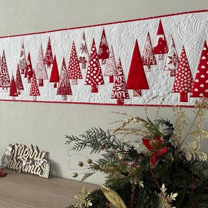May include: A Christmas-themed wall hanging featuring red and white quilted Christmas trees. The trees have various patterns, including polka dots and text. The border is red, and the background is white with quilted patterns. A wooden sign that says "Merry Christmas" is on the table below.
