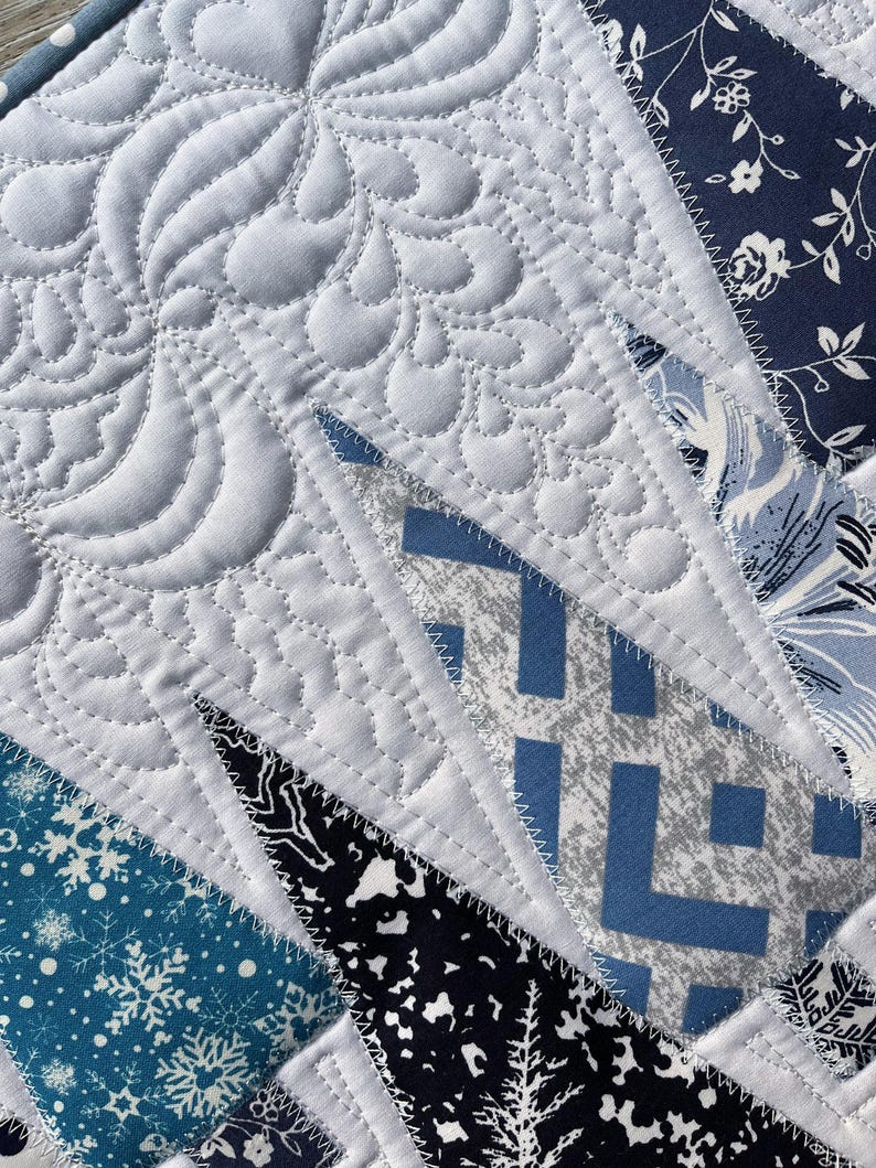 May include: A patchwork quilt featuring a variety of blue and white fabric patterns, including snowflakes, floral prints, and geometric shapes. The textile is finished with intricate white thread quilting in swirling, organic, and wavy patterns.