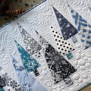 May include: A quilted table runner featuring a winter theme with various triangular fabric trees in shades of navy, light blue, and white. The background fabric is white with intricate swirling and circular machine-stitched quilting patterns.
