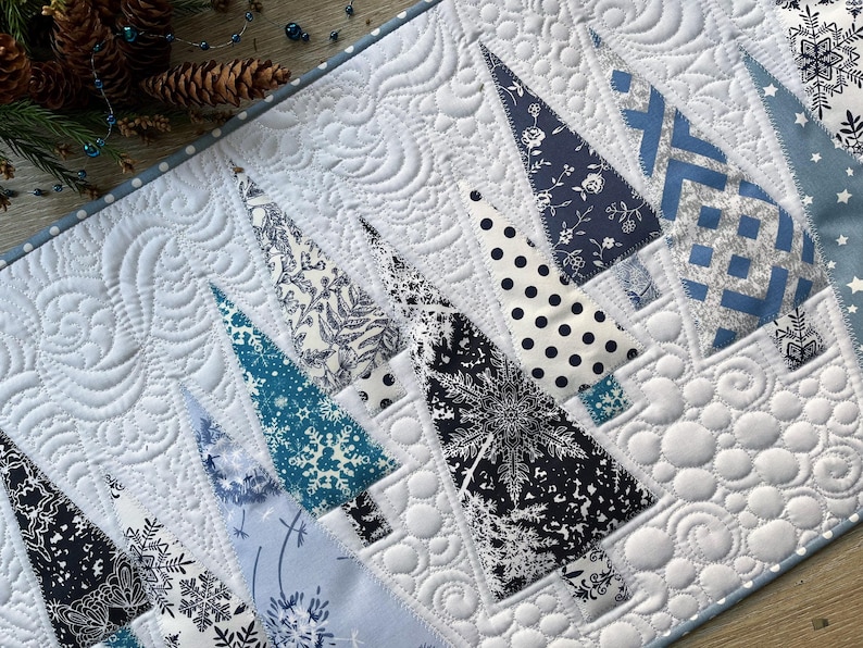 May include: A quilted table runner featuring a winter theme with various triangular fabric trees in shades of navy, light blue, and white. The background fabric is white with intricate swirling and circular machine-stitched quilting patterns.