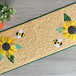 Puede incluir: Un camino de mesa acolchado rectangular de tela color canela claro con un intrincado patrón floral en espiral. Presenta dos grandes girasoles con pétalos amarillos y centros de lunares blancos y negros, acentuados por hojas verdes y dos pequeñas abejas.