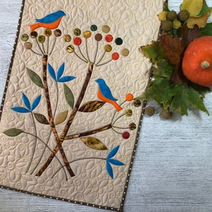 May include: A quilted table runner with a cream background and a brown polka dot border. The runner features a design of blue and orange birds perched on branches with colorful berries and leaves. A small pumpkin and fall foliage are on the side.