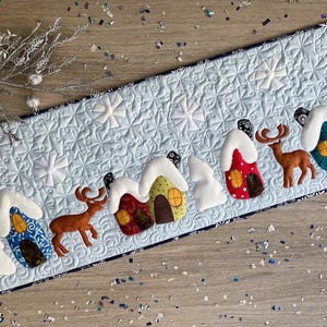May include: A light blue quilted table runner with a winter village scene. The runner features appliquéd houses with snow-covered roofs, snowflakes, and reindeer. The houses are in various colours, including red, blue, and yellow.