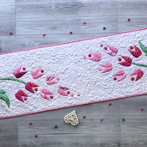 May include: A quilted table runner featuring a floral design with pink and red tulip-like shapes on a white background. The runner has a pink border and is decorated with green leaves. Decorative hearts and a butterfly are placed around the runner.