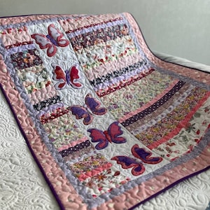 Könnte beinhalten: Handgefertigte Patchwork-Quilt mit verschiedenen floralen Mustern in Rosa, Lila und Weiß. Applikationen von Schmetterlingen in Rosa- und Lilatönen sind auf dem Quilt verteilt. Der Quilt hat einen rosafarbenen Rand mit einer violetten Einfassung.