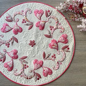 Peut inclure: Un tapis en tissu matelassé rond avec un fond crème et des surpiqûres complexes. Le motif présente une disposition circulaire de cœurs à motifs roses et rouges, des volutes en forme de vigne et de petites feuilles, bordé d'un liseré rouge.