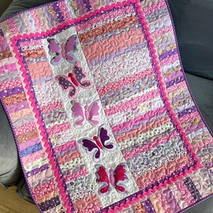 May include: A rectangular quilt featuring a central vertical panel with five appliquéd butterflies in shades of pink and purple. The quilt is bordered by a bright pink rickrack trim and alternating vertical fabric strips in floral, polka dot, and star patterns in various pink, purple, and white hues.