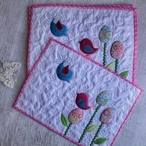May include: Two white quilted placemats with pink polka dot borders. Each mat features embroidered birds and Easter egg designs in shades of blue, pink, and green. The placemats are decorated with floral patterns.
