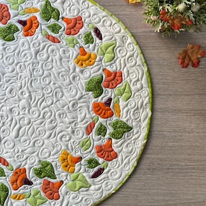Könnte beinhalten: Eine runde, gesteppte Stoffmatte mit weißem Hintergrund und komplizierten, wirbelnden Steppmustern. Die Matte ist mit einer Applikationsbordüre aus orangefarbenen und gelben Blumen sowie grünen Blättern verziert und hat einen grünen Rand.