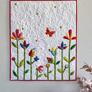 Pode incluir: Uma tapeçaria de parede acolchoada com um design floral colorido. O fundo branco é adornado com costuras intrincadas e pequenos detalhes verdes. As flores são de várias cores, com uma borda vermelha.