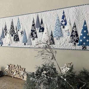 May include: A quilted Christmas tree table runner with blue and white patterned trees. The trees are set against a white background with decorative stitching and a grey border. The words "Merry Christmas" are visible in the foreground.