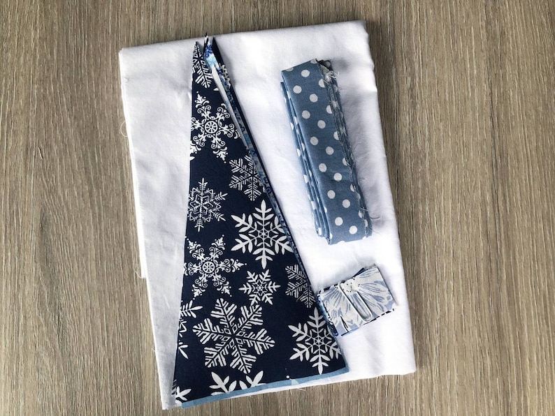 May include: A sewing kit featuring a large white fabric square, a triangular piece of dark navy blue fabric with a white snowflake pattern, a folded strip of light blue fabric with white polka dots, and a small rectangular piece of floral fabric.