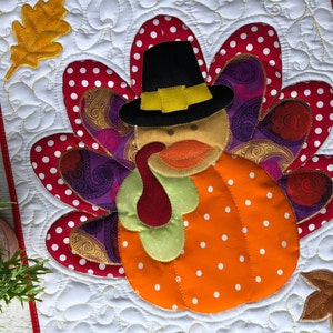 Può includere: Un arazzo trapuntato rettangolare con un'applicazione centrale a forma di tacchino con corpo a zucca, cappello da pellegrino nero e piume colorate con motivi a pois rossi e paisley viola, accentuato da due foglie in tessuto su fondo bianco.