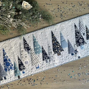 May include: A rectangular quilted table runner featuring a repeating pattern of stylized Christmas trees. The trees are various shades of blue and white, with different patterns. The background is white with a quilted design. Decorative stars and greenery are in the background.