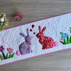 May include: A white quilted Easter table runner with a pink border. It features two appliqued bunnies, one purple and one red, with floral patterns. Hearts and flowers decorate the runner, with a small floral arrangement and a butterfly.