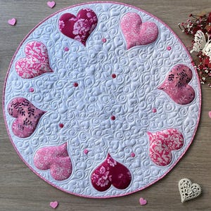 May include: A round, white quilted placemat with a pink border. It features appliqued pink hearts with floral patterns and small pink dots. The background has a swirling pattern. Decorative hearts and a lace heart are scattered around.