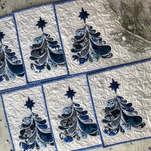 Puede incluir: Un juego de manteles individuales y un camino de mesa navideños acolchados. Cada mantel individual presenta un diseño estilizado de árbol de Navidad en tonos azules y blancos, coronado por una estrella azul. Los manteles y el camino tienen un fondo blanco con un borde azul.