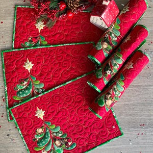 May include: A set of rectangular quilted placemats in vibrant red fabric featuring a green Christmas tree motif with gold star accents and green trim. The surface has a swirling stitched pattern, and the set includes matching rolled fabric napkins.