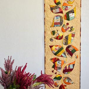 May include: A quilted wall hanging featuring a stylized leaf design. The leaves are composed of various fabric scraps in shades of orange, green, yellow, and red. The background is a light tan color with a quilted pattern.