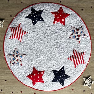 May include: A white quilted table runner with a red, white, and blue star design. The runner is circular and has a scalloped edge.