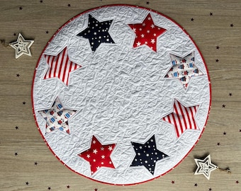 May include: A white quilted table runner with a red, white, and blue star design. The runner is circular and has a scalloped edge.