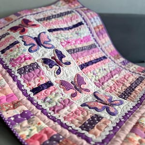 Könnte beinhalten: Eine handgefertigte Steppdecke mit einem Patchwork-Design in Rosa-, Lila- und Weißtönen. Die Steppdecke ist mit applizierten Schmetterlingsmotiven verziert. Die Kanten sind mit einem gewellten Rand versehen.