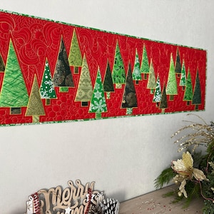 May include: A rectangular quilted table runner featuring a vibrant red background with a swirling stitched pattern. The runner displays a variety of green Christmas tree appliques in different shades, textures, and patterns, all bordered by a green trim.