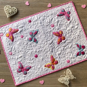 May include: A white quilted placemat with pink trim, featuring embroidered butterflies in shades of pink, yellow, and blue. The placemat has a detailed floral pattern and is surrounded by pink heart-shaped decorations.
