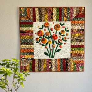 May include: A handmade quilt with a floral design. The central square features a bouquet of orange, red, and yellow flowers with green stems and leaves on a white background. The border is made of colourful fabric strips.