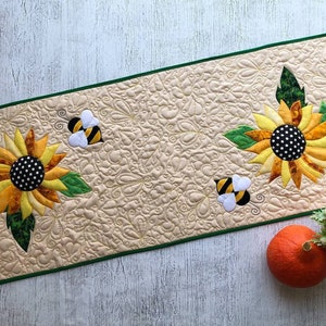 May include: A quilted table runner featuring appliqued sunflowers with yellow, orange, and black centers, and green leaves. Two appliqued bees are also present. The runner has a beige background with a green border.
