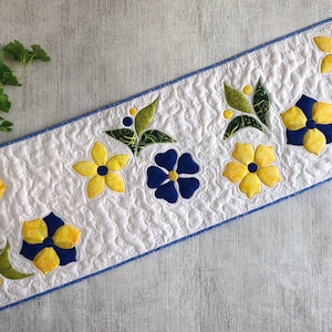 May include: A white quilted table runner with a blue border, adorned with appliqued yellow and blue flowers, and green leaves. The floral design is evenly spaced across the runner's length, creating a decorative accent for any table setting.