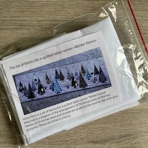 May include: A fabric kit for a quilted table runner titled Winter forest, enclosed in a clear plastic bag. The kit includes a printed instruction sheet featuring a diagram of various patterned Christmas tree shapes on a white background.