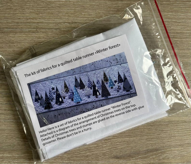 May include: A fabric kit for a quilted table runner titled Winter forest, enclosed in a clear plastic bag. The kit includes a printed instruction sheet featuring a diagram of various patterned Christmas tree shapes on a white background.