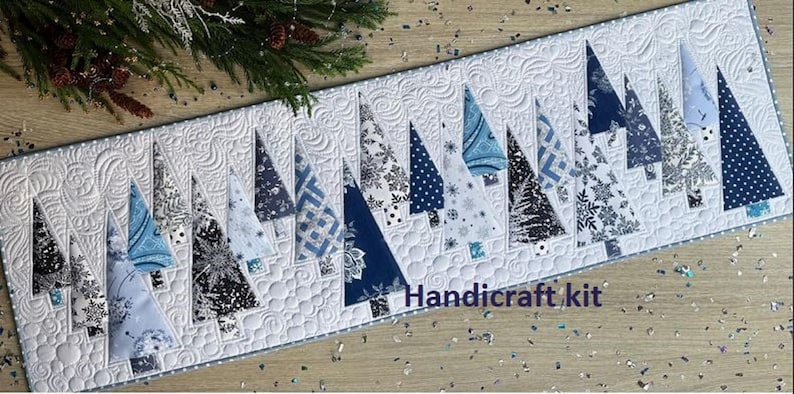 May include: A rectangular quilted table runner featuring a white background with intricate swirling patterns and a series of triangular fabric Christmas trees in various shades of blue, navy, and white. The text Handi-craft kit is visible.