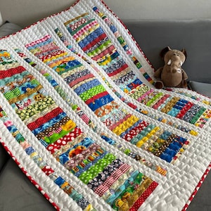 May include: A white baby quilt with a patchwork design featuring colourful fabric strips with various patterns, including polka dots, stripes and floral prints. The quilt has a white border with red polka dots.