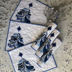 Pode incluir: Um conjunto de jogos americanos acolchoados e guardanapos enrolados a condizer com fundo branco, costuras intrincadas e um padrão de árvore de Natal azul, rematado com uma estrela azul-escura. As bordas têm um acabamento em viés azul.