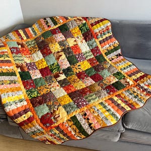 Könnte beinhalten: Eine Patchwork-Decke mit einem Gitter aus quadratischen Stoffstücken in Herbstfarben wie Orange, Gelb, Rot, Grün und Braun. Die Decke hat einen Rand aus vertikalen Stoffstreifen und eine dunkelbraune Einfassung sowie verschiedene Blumen- und Blattmuster.